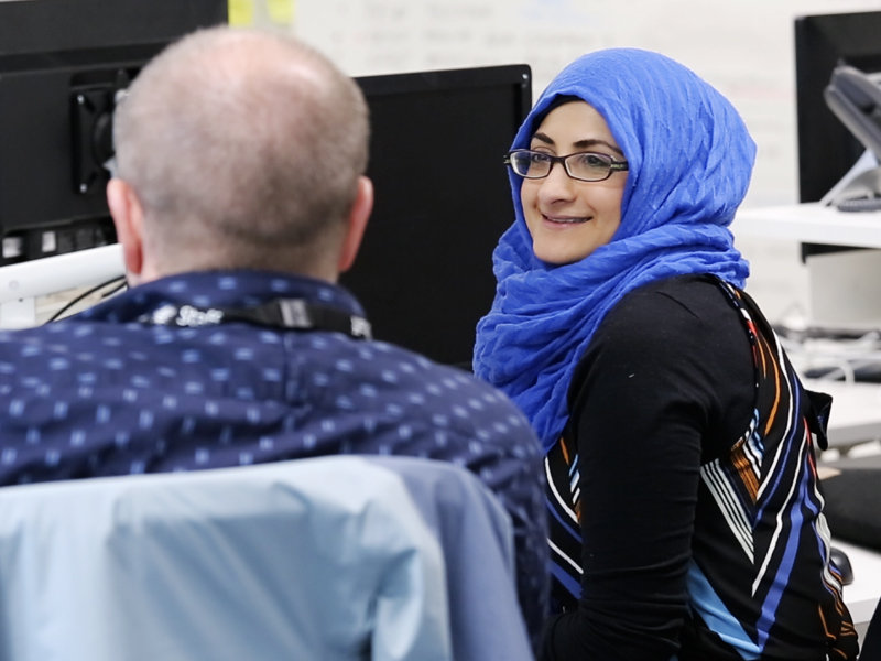 A woman wearing a hijab smiling and talking to a colleague. They are seated in front of computer screens.