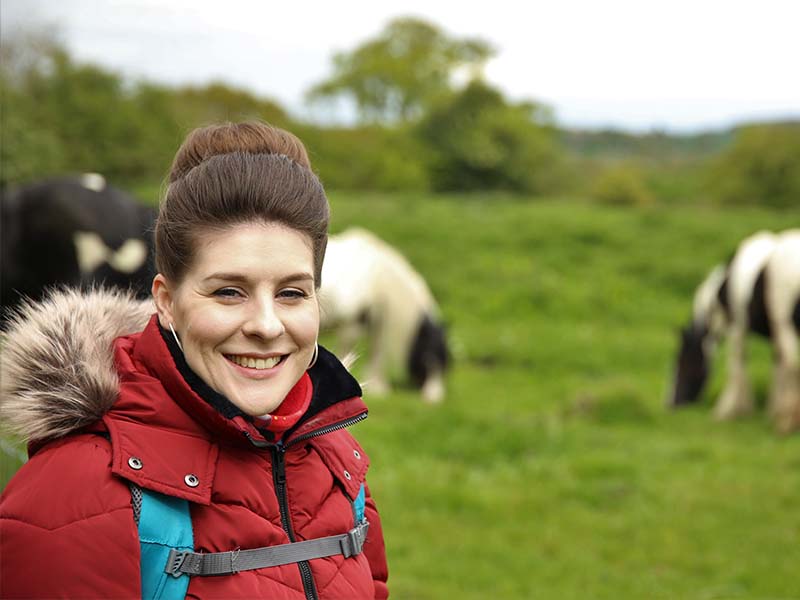 Michelle, wearing a red coat, standing in a field with some horses