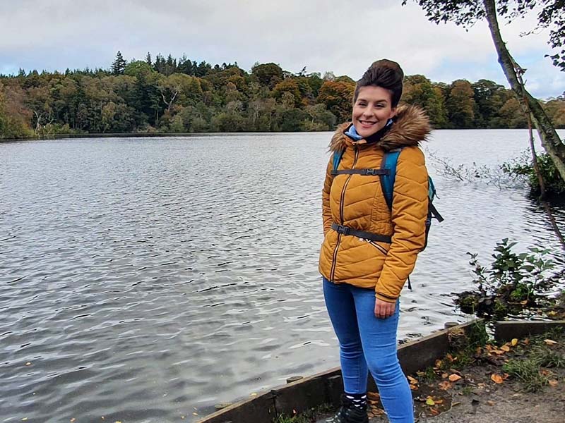 Michelle, wearing a yellow coat, standing in front of a lake