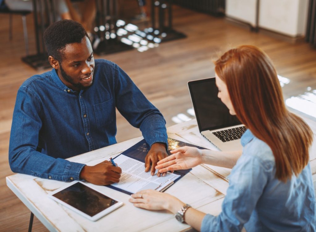 Two people sit across a desk from each and smile. There are papers and devices in front of them.