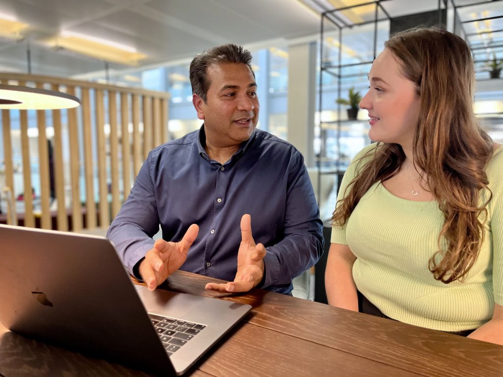 Parag and Emma, two DWP Digital colleagues, sit at a desk in an open plan office. They are talking, with an open laptop computer in front of them.