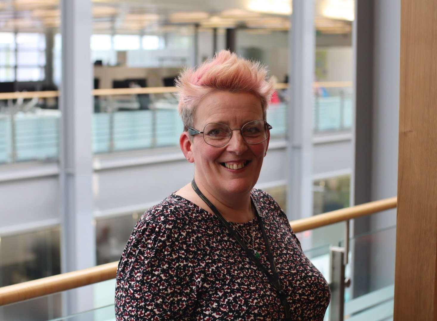 Roni, a woman with short, colourful hair and glasses, smiling towards the camera. She is standing in a large open plan office.