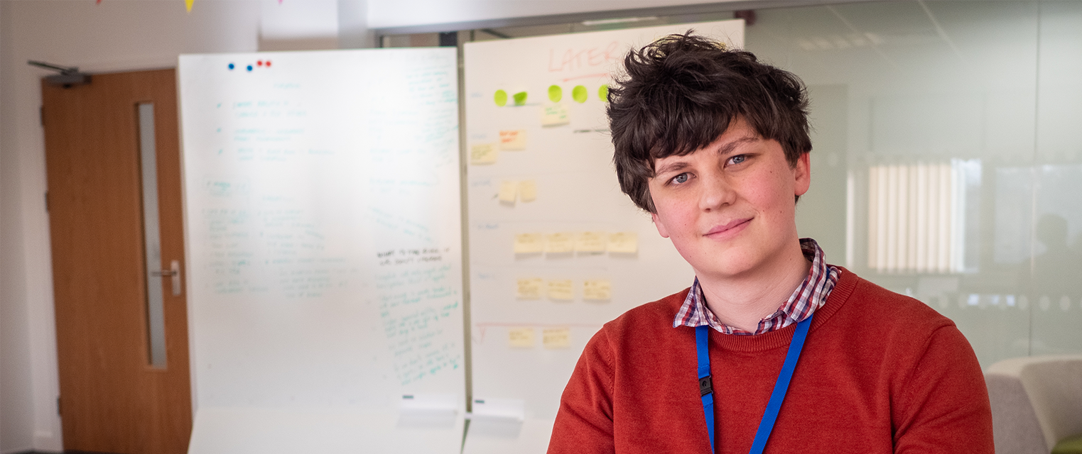 Luke smiles at the camera while sitting in a brightly lit office. He is wearing a red jumper. There is a whiteboard behind him.