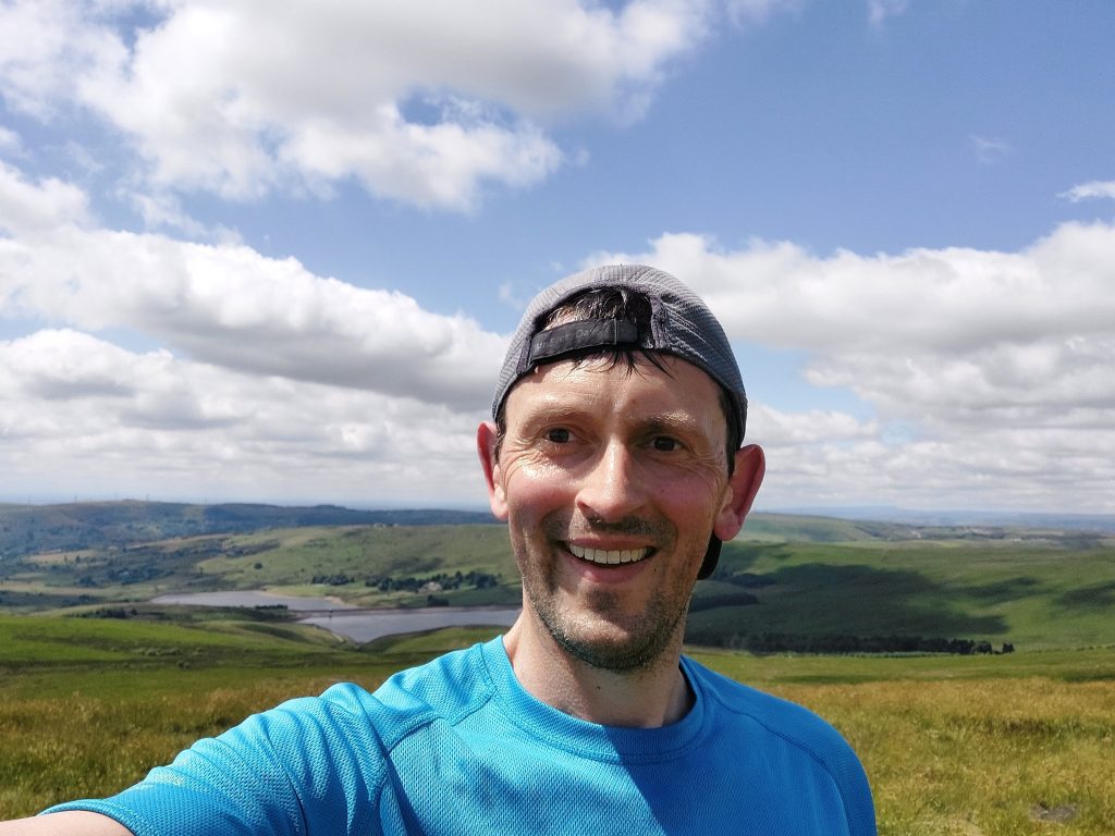 Andy Tyack smiling at the camera on a sunny day. Behind him are hills and a reservoir.