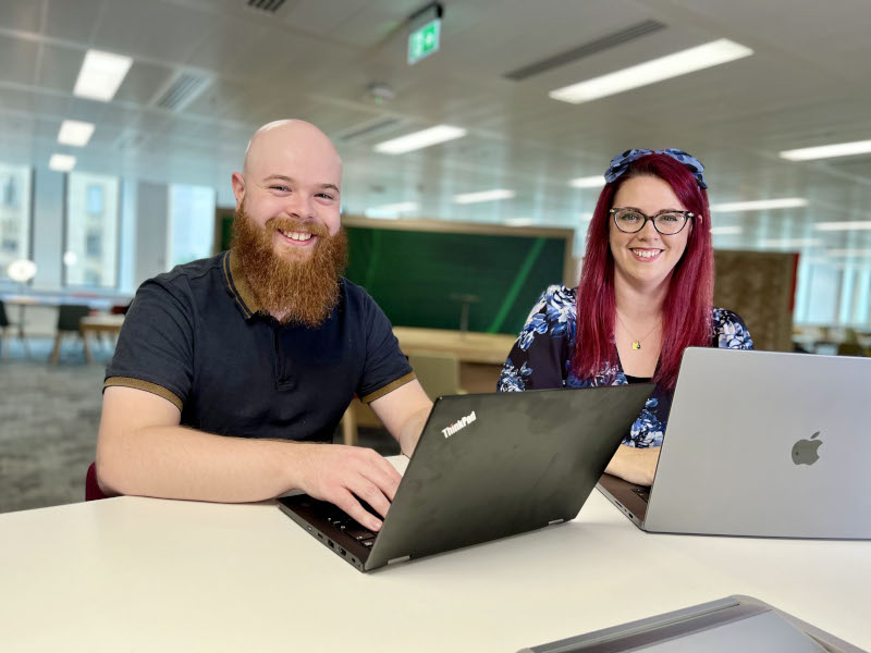 Armand has a long beard and is wearing a blue shirt. Hannah has red hair and is wearing a blue print dress. They smile at the camera, with laptops on the desk in front of them.