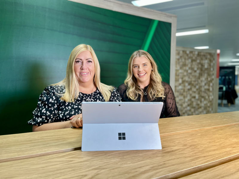 Jacqui is wearing a blue print dress, and Catherine wears a black top. They are sitting next to each other and smiling at the camera, with a laptop in front of them.