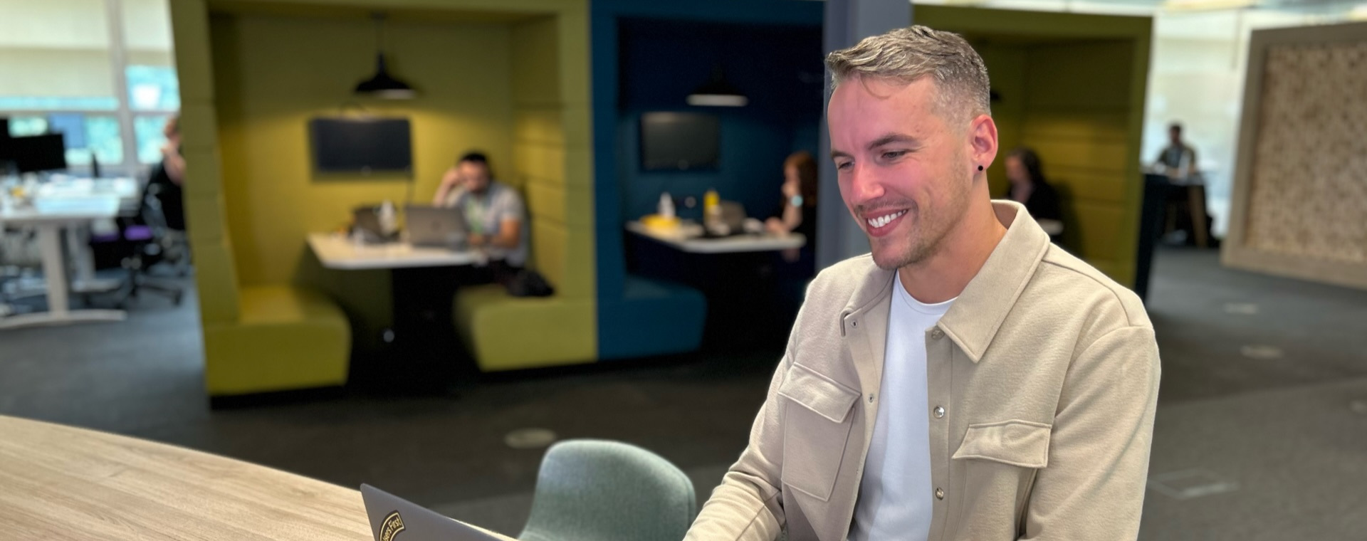 Max smiles as he sits at a desk in an open plan office and works on a laptop computer