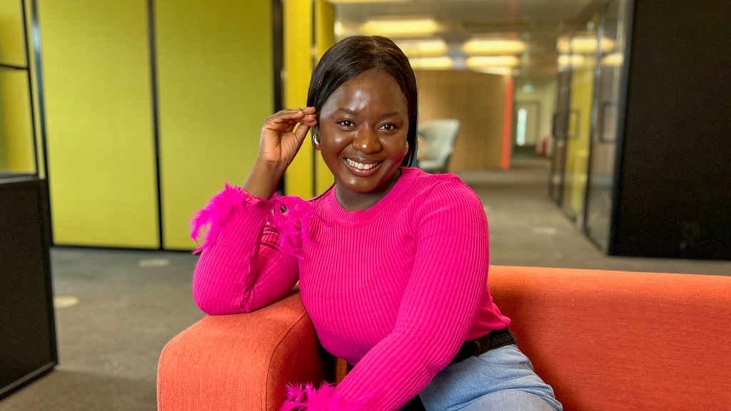 Olu is wearing a bright pink jumper and smiling. She is sitting on an orange sofa.