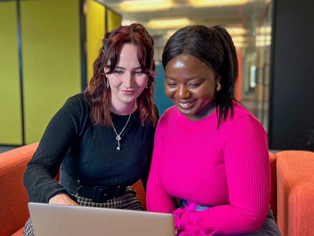 Two women sit together on a sofa, and smile as they work together on a laptop