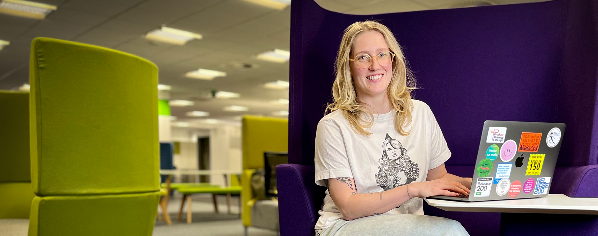 Kathryn sits in an open office meeting space. She has a laptop open in front of her with stickers on it, and she is smiling.