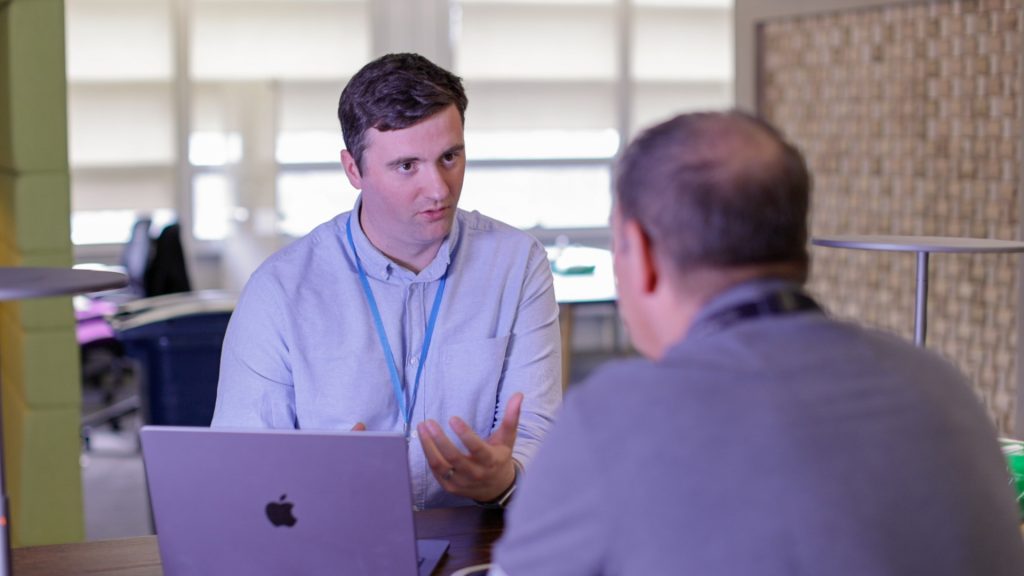 Man sat with colleague providing advice and signalling to a laptop.